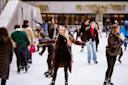 People enjoying ice skating on a rink, bundled up in winter clothing. In the center, a person with long hair and a plaid coat joyfully holds out their arms, surrounded by other skaters. The background features a building and some trees.