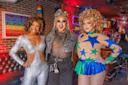 Three drag performers pose inside a colorful bar with brick walls. One wears a sparkling silver jumpsuit, one wears a sheer top with bold makeup, and one wears a curly wig with a blue and green star-patterned outfit.