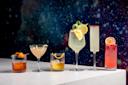 Five elegantly presented cocktails are lined up on a white surface against a starry background. Each drink is uniquely garnished with items like orange peel, lime, and herbs, showcasing a variety of colors and glass styles.