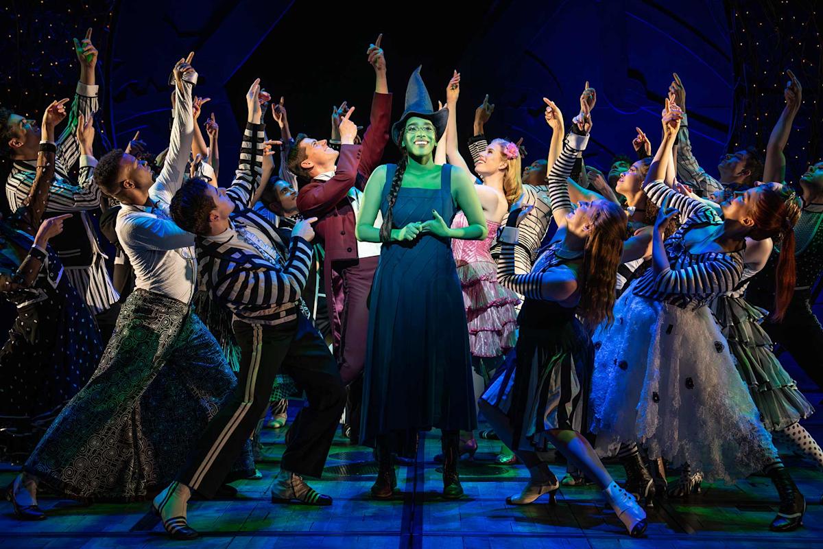 A group of theater performers in colorful costumes surround a smiling woman with green skin, who stands center stage with her arms slightly raised, as others point and look toward her under bright stage lights.