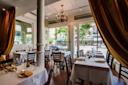 Elegant restaurant interior with white tablecloths, wooden chairs, and gold curtains. Large windows let in natural light and show a view of a city street with trees, cars, and storefronts outside. A chandelier hangs from the ceiling.