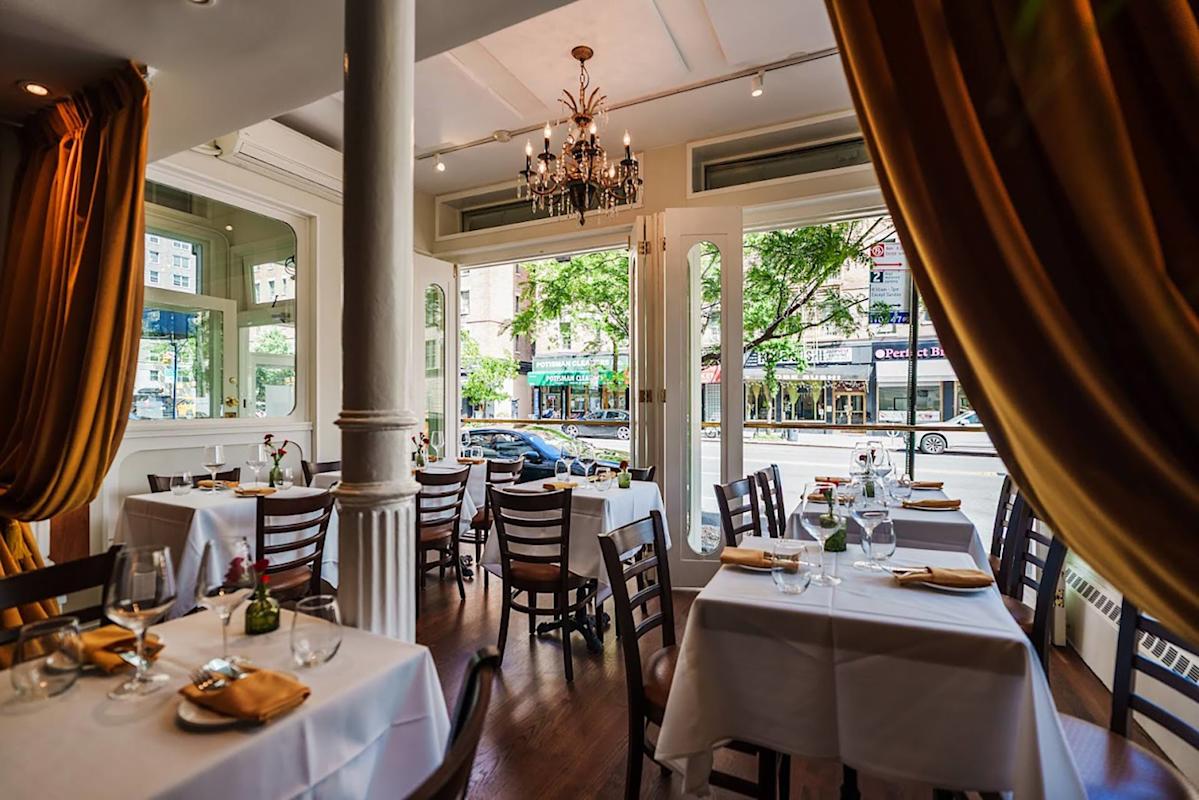 Elegant restaurant interior with white tablecloths, wooden chairs, and gold curtains. Large windows let in natural light and show a view of a city street with trees, cars, and storefronts outside. A chandelier hangs from the ceiling.