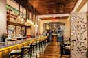 Stylish restaurant interior with a long bar lined with high chairs, hanging glass pendant lights, wooden floors and ceiling, decorative screen panels, and shelves of bottles behind the bar.