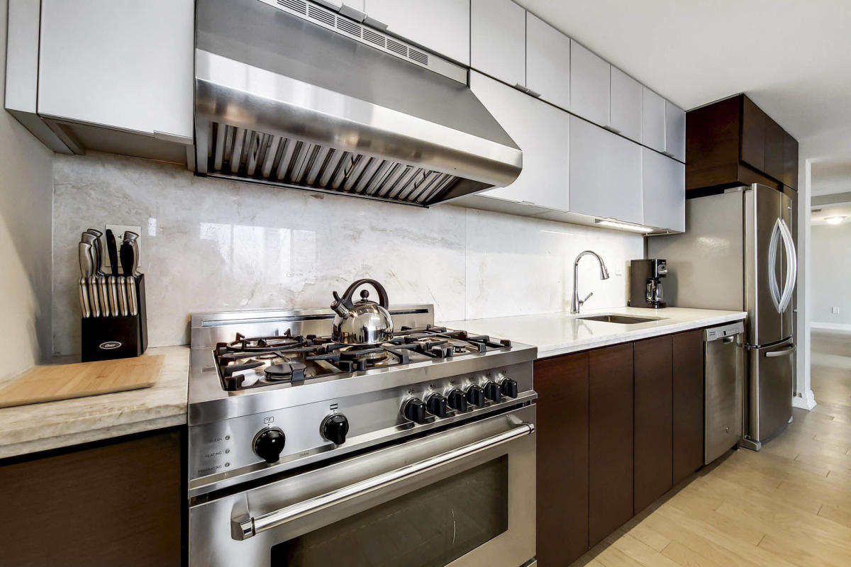 Modern kitchen with stainless steel appliances, including a large gas stove and range hood. A silver kettle sits on the stove. White and dark wood cabinets line the walls, and a set of knives rests on a wooden block. Light-colored flooring throughout.