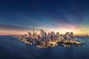 Aerial view of a city skyline at dusk, with skyscrapers illuminated and a mix of deep blue and orange hues in the sky. The water surrounding the city reflects the lights and colors, creating a serene and vibrant urban scene.