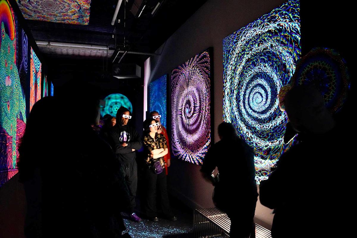 A group of people wearing 3D glasses view vibrant, swirling psychedelic art displayed on dark gallery walls, illuminated by blacklight. The scene has a colorful, immersive, and futuristic atmosphere.
