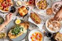An overhead view of a table filled with various dishes including a bread roll, hot dog topped with slaw, fried fish, sliced meats, colorful salads, and glasses of red and white wine. Hands reach in to serve and enjoy the meal.