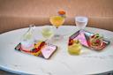 A white table with five elegant cocktails, each on a metallic tray. Garnishes include olives, flowers, red peppers, and citrus. Some drinks come with snacks like crackers and caviar. The background features a striped fabric.