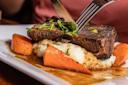 A person cuts into a piece of braised beef short rib served over mashed potatoes with roasted carrots, garnished with chopped parsley and drizzled with brown gravy on a white plate.