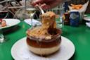 A hand lifts gooey, melted cheese from a bowl of French onion soup on a white plate, with wine glasses, fries, and other dishes on a green table in the background.
