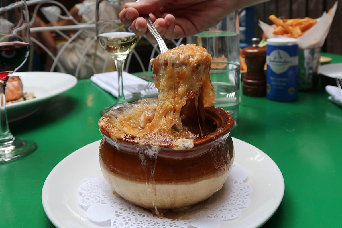 A hand lifts gooey, melted cheese from a bowl of French onion soup on a white plate, with wine glasses, fries, and other dishes on a green table in the background.