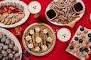 A variety of desserts on a red table, including a large cookie cake with whipped cream, churros with chocolate sauce, powdered donuts, chocolate truffles, and a plate of cookies with strawberries. Some drinks are also visible.