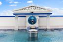 A decorative water fountain with a blue lion head spout set in a white, Greek-style structure above a pool. Sculptures and columns adorn the top, with steam rising from the water and a large metal container in front.