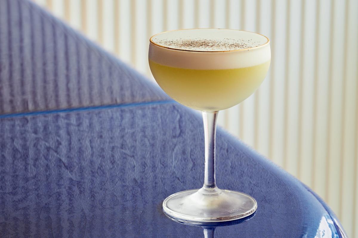 A coupe glass filled with a yellow cocktail topped with a thick layer of foam and sprinkled with dark powder, placed on a glossy blue surface.