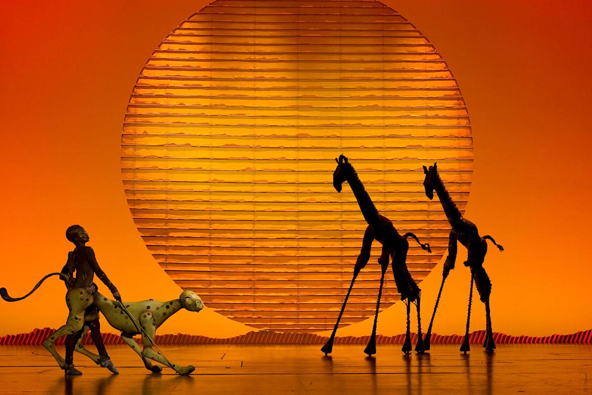 Silhouetted puppets of a cheetah, human, and giraffes are staged against a large, glowing orange sun, evoking an African savanna at sunset.