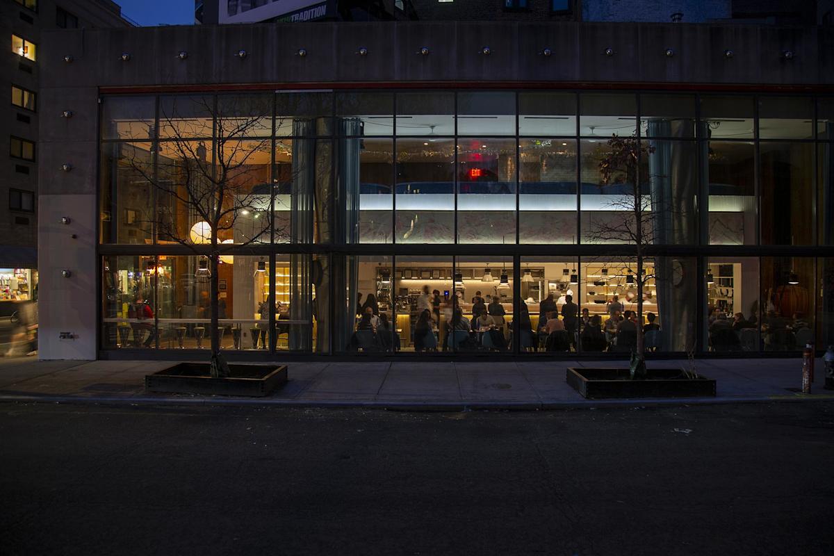 A restaurant with floor-to-ceiling glass windows shows people dining inside at dusk, with warm lights illuminating the interior. The street outside is empty, with planters and bare trees in front of the building.
