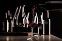 Red wine is being poured from a bottle into a glass on a dark table, with a decanter, more wine glasses, and bottles blurred in the background, creating an elegant and moody atmosphere.
