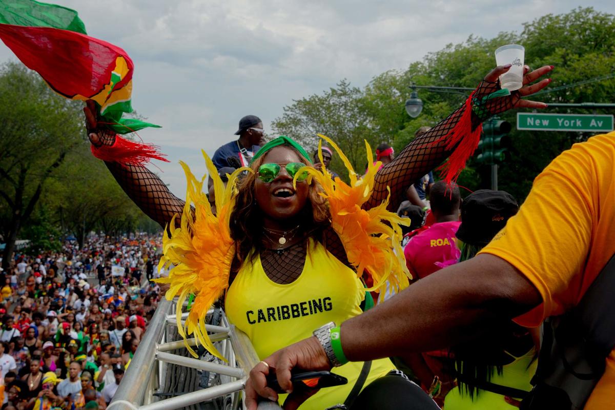 A woman in a bright costume with yellow and orange feathers joyfully waves a flag at a lively outdoor parade. She wears sunglasses and a “CARIBBEING” shirt. A large crowd and a “New York Av” street sign are visible.