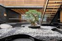 A modern indoor space featuring a bonsai tree in a planter, surrounded by smooth gray pebbles and shallow black water pools, with a floating staircase and wooden ceiling above.