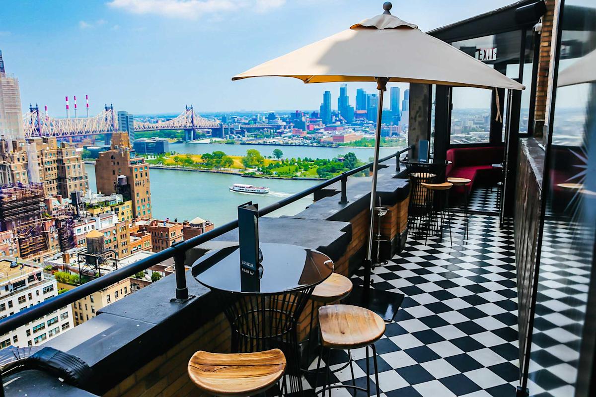 Rooftop bar with black and white checkered floor, round tables, wooden stools, and a large umbrella overlooking a cityscape, river, bridge, and distant skyline on a sunny day.