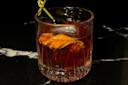 A glass of dark cocktail with ice, garnished with orange peel and a skewer holding two black cherries, sits on a black marble surface.