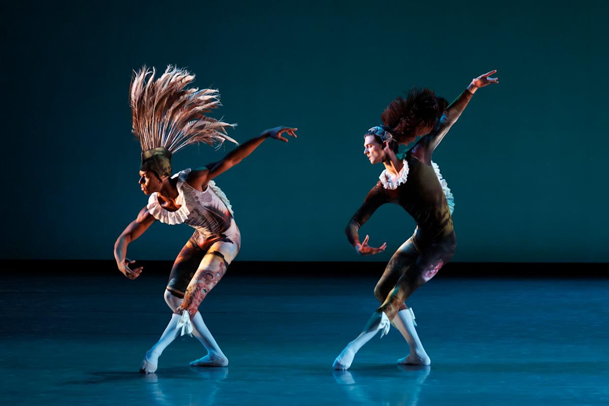 Two ballet dancers in colorful costumes and headpieces pose dramatically on stage, extending their arms and legs with expressive, fluid motion against a blue-lit background.