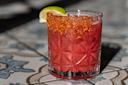 A cocktail with a deep red color is served in a textured glass. The rim is coated with a spicy red seasoning, and a lime wedge is placed on the rim. The glass is on a patterned surface.