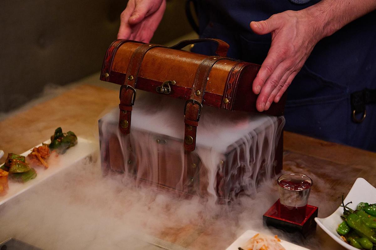 A person opens a vintage wooden chest with metal clasps, releasing dramatic white smoke that spills over the sides. Plates of food and a glass of liquid sit on the wooden table nearby.