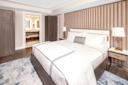 Modern hotel room with a large bed featuring white linens and a grey throw pillow. Wooden headboard and bedside tables with lamps. Open door reveals a bathroom with a double sink vanity. A plush blue and gray rug lies on the wood floor.