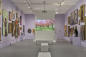 A gallery with walls covered in various framed artworks. At the center is a projection screen showing a vibrant image of a cherry blossom park. Sculptures are placed on pedestals around the room. The floor is gray, and the walls are lavender.