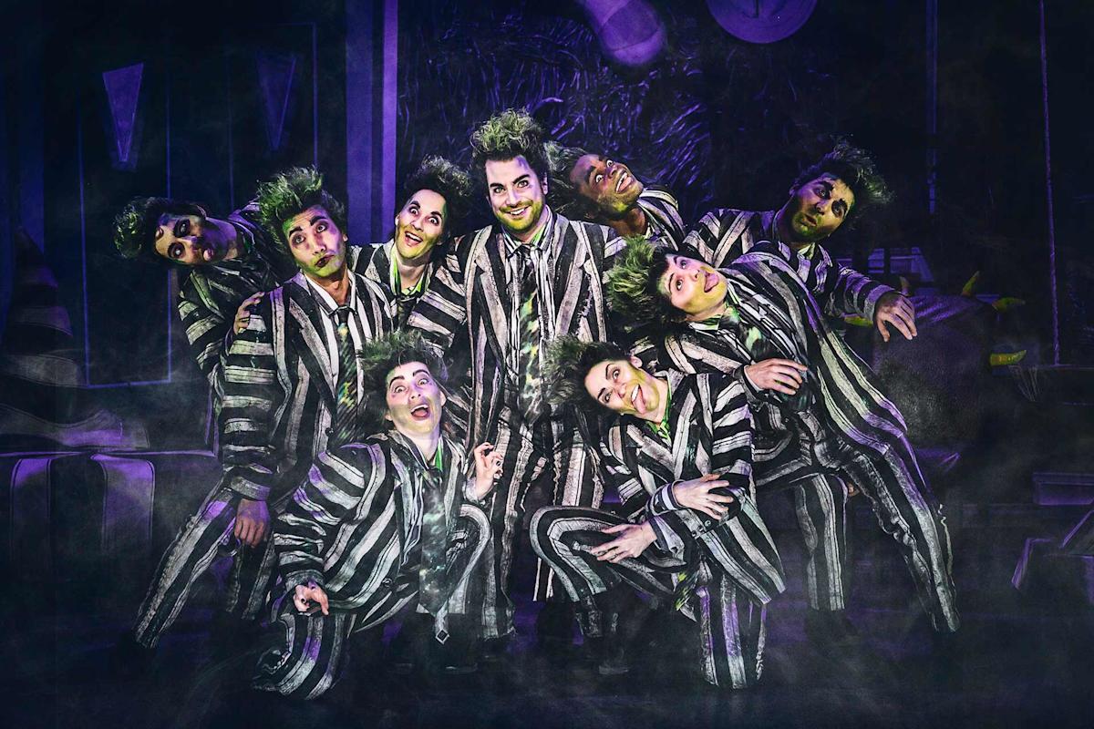 A group of actors in black-and-white striped costumes and green-tinted makeup pose together onstage, smiling and making exaggerated faces, in a theatrical, foggy setting with dramatic lighting.