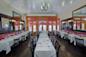 Elegant restaurant interior with white tablecloths and dark wooden chairs. A chandelier hangs from the ceiling, and large mirrors decorate the walls. Tables are set for dining, with neatly arranged plates, glasses, and silverware.