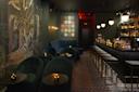 A dimly lit, elegant bar with velvet seating, a long counter with high stools, ornate wall art, and pendant lights. The floor has a geometric pattern, and the atmosphere is cozy and sophisticated.