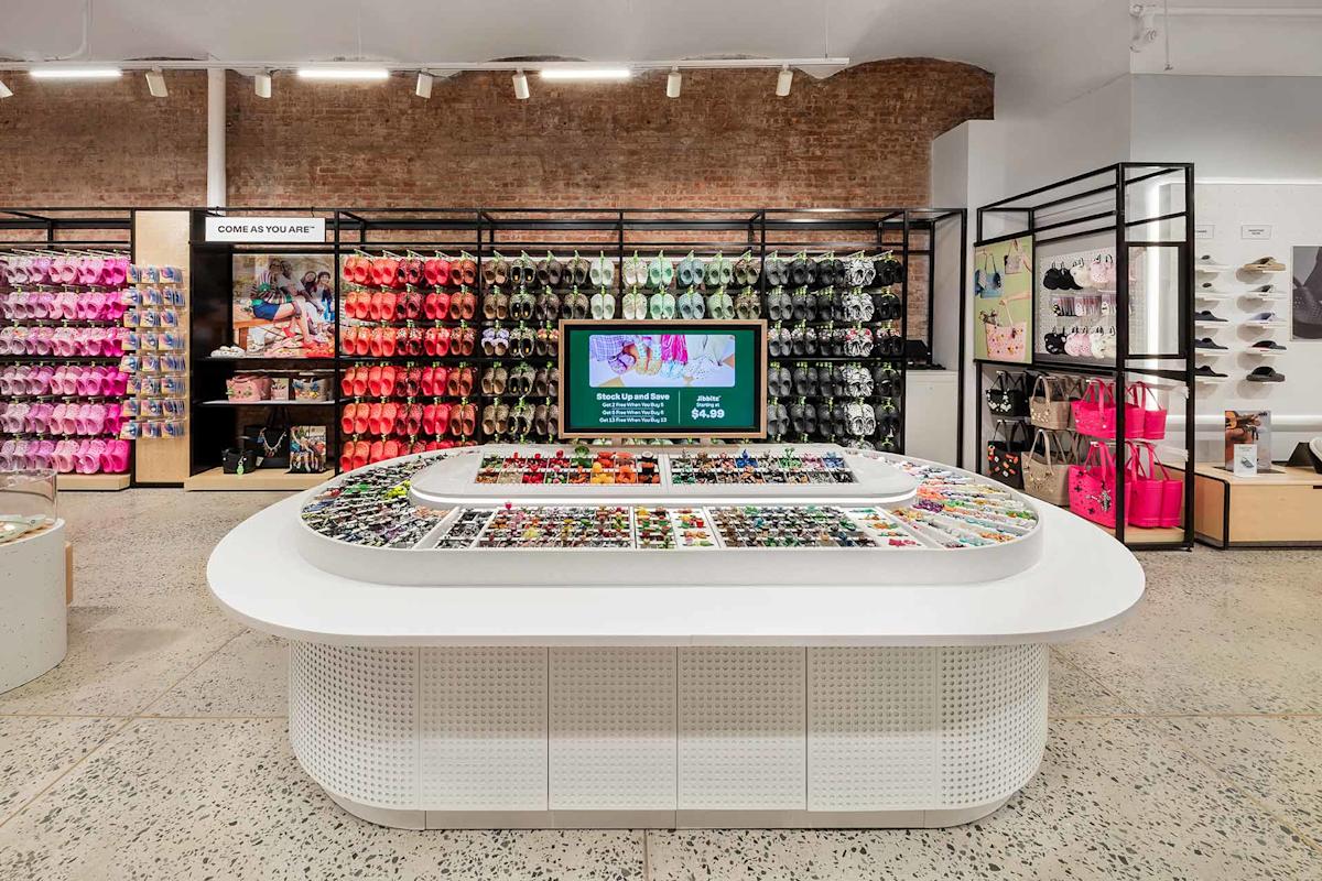 A brightly lit retail store with colorful footwear and bags displayed on wall racks, a central white display table filled with accessories, and a digital screen showcasing promotions. Brick and white walls create a modern aesthetic.
