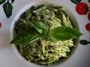 A plate of pasta coated in green pesto sauce, garnished with fresh basil leaves and grated cheese. The plate has tomato and basil leaf decorations on the rim.