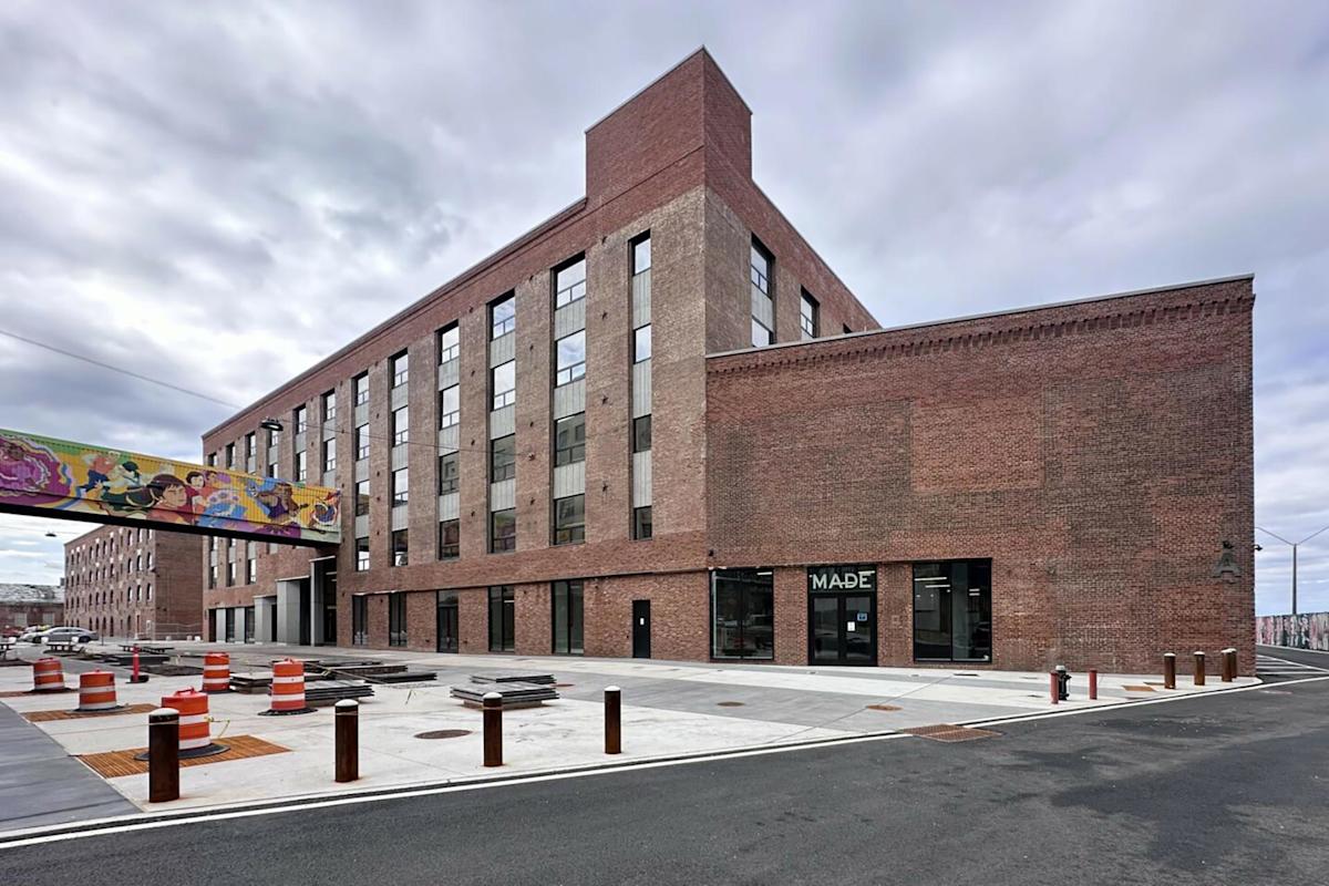 A large brick industrial building with tall windows and a sign reading "MADE" on the ground floor. A colorful mural decorates a pedestrian bridge. Orange traffic barrels and construction materials are visible on the wide sidewalk.