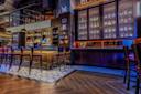 A stylish bar interior with high wooden stools lining a bar counter, illuminated by pendant lights. Shelves stocked with various bottles are in the background. The floor has hexagonal tiles and wood, and the atmosphere is cozy and inviting.