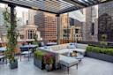 A modern rooftop lounge with casual seating, tables, and potted plants. The area overlooks tall city buildings.