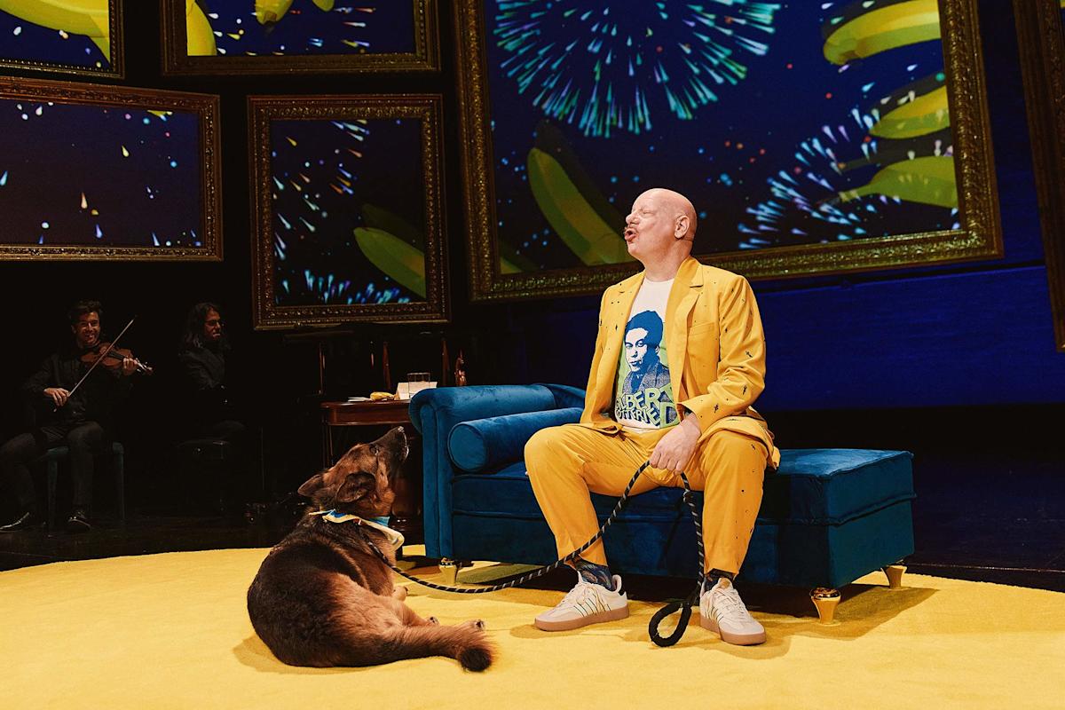 A bald man in a bright yellow suit sits on a blue couch on stage with a dog at his feet. Behind him are large screens displaying bananas and fireworks. Musicians are partially visible in the background.