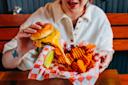 A person holding a cheesy burger with melted cheese stretching, next to waffle fries, a pickle spear, and a cup of ketchup in a red-and-white checkered basket.