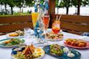 A table set outdoors with assorted brunch dishes, including avocado toast, salad, eggs, pastries, and colorful drinks in flutes and glasses, decorated with artificial blue butterflies, greenery visible in the background.