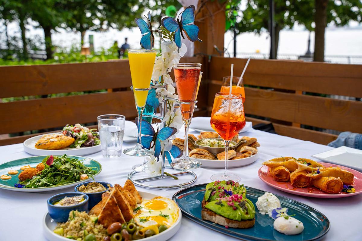 A table set outdoors with assorted brunch dishes, including avocado toast, salad, eggs, pastries, and colorful drinks in flutes and glasses, decorated with artificial blue butterflies, greenery visible in the background.