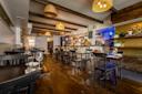 Cozy restaurant interior with wooden flooring and beams. Tables are set with glassware and napkins. Wicker pendant lights hang from the ceiling. The bar area is in the background, adorned with shelves and decorative items.
