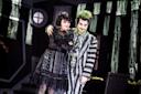 Two actors in theatrical costumes stand together onstage. One wears a black lace dress and curly dark hair; the other has green hair and face paint, wearing a striped suit. The set behind them has a spooky, gothic style.