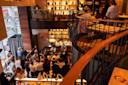 A stylish, modern restaurant with two levels, crowded with people enjoying food and drinks. Warm lighting, shelves with decorative items, and a busy bar area create a lively atmosphere.