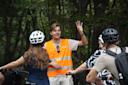 A man wearing an orange reflective vest gestures with his hand while talking to a group of cyclists, who are wearing helmets, in a wooded outdoor setting.