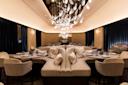 Elegant restaurant interior with plush seating, round tables set with glassware, and decorative cushions. A striking chandelier with teardrop-shaped lights hangs from the ceiling. The ambiance is warm, with soft lighting and dark drapes lining the walls.
