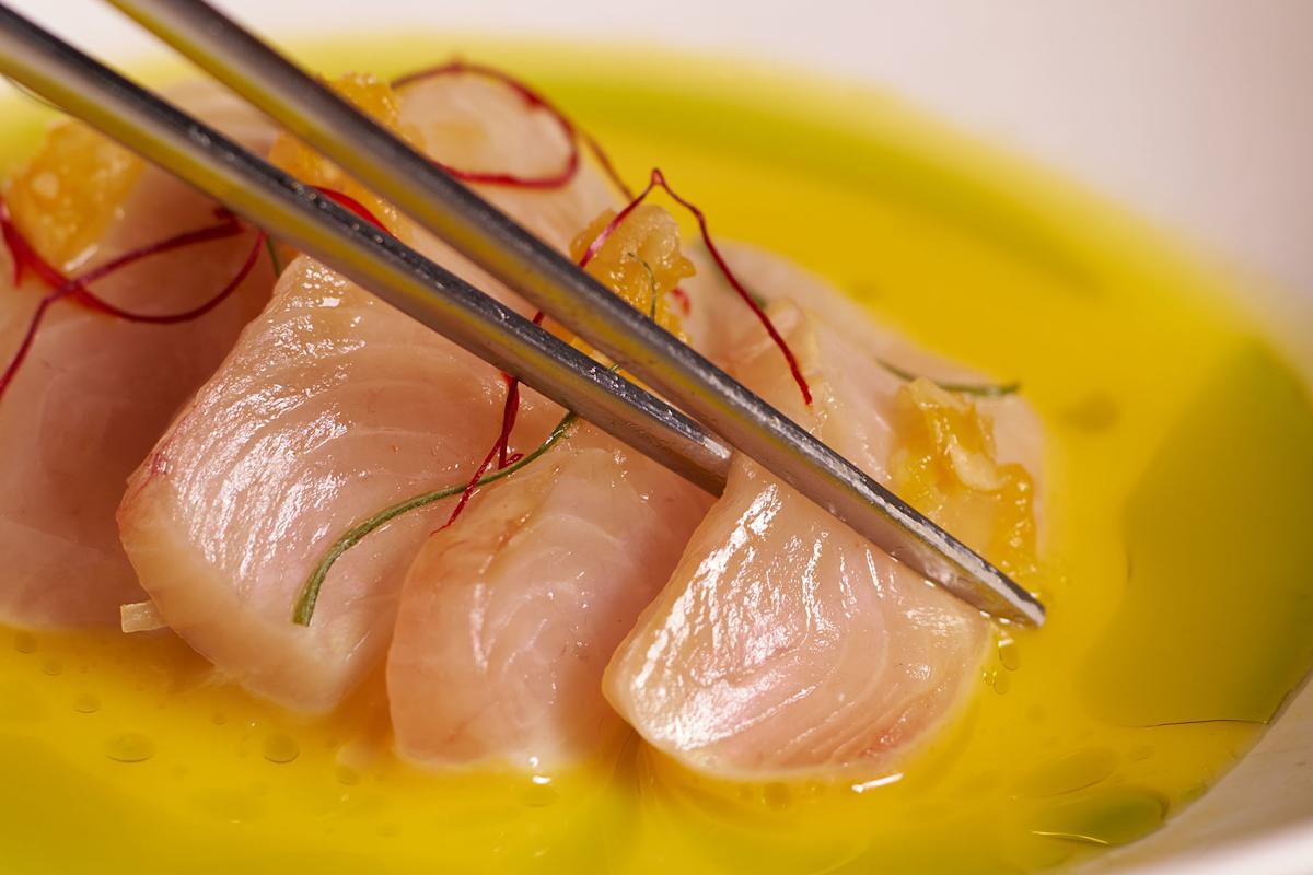 Close-up of metal chopsticks holding a slice of raw fish atop several pieces, garnished with thin red strands and served in a yellow oil-based sauce.