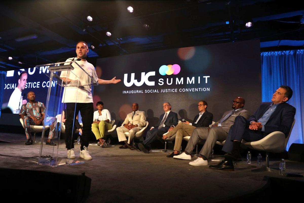 A speaker stands at a podium addressing an audience at the WJC Summit, with seven seated panelists on stage in front of a large screen that reads “WJC Summit Inaugural Social Justice Convention.”.
