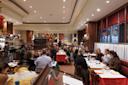 A lively restaurant scene with people dining at tables, chatting, and enjoying drinks. The interior features warm lighting, wooden details, large windows, and floral centerpieces adding color to the elegant setting.